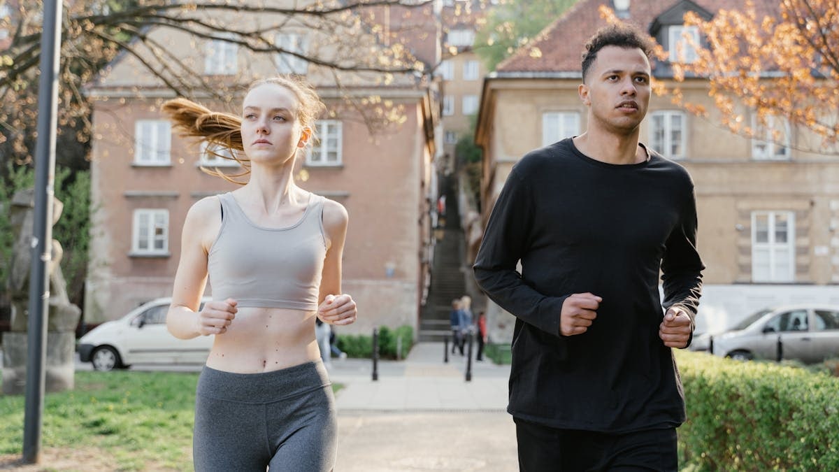 A young woman and man jogging on a city street during the day, wearing sportswear.